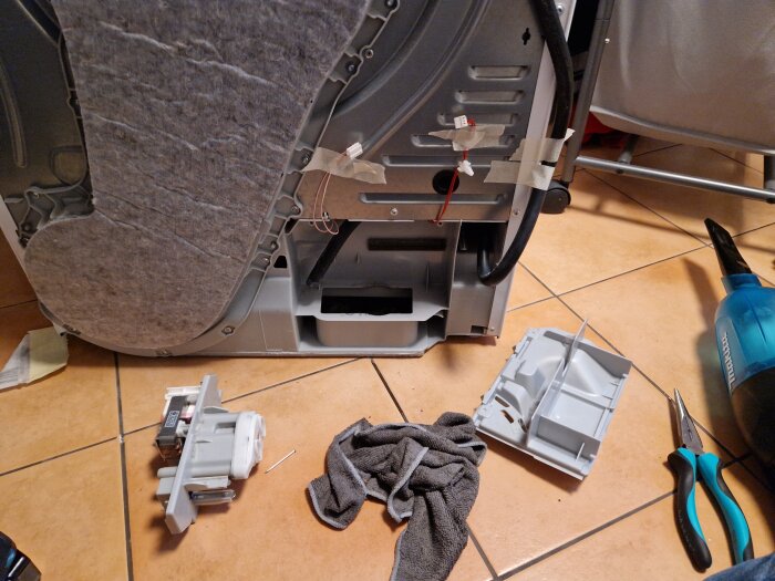 Demonterad tvättmaskinsdelar på golv med verktyg och duk. Reparation eller service verkar pågå.