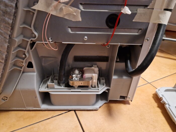 Insida av en apparat, troligen en tvättmaskin, med exponerade komponenter och sladdar, på ett golv.