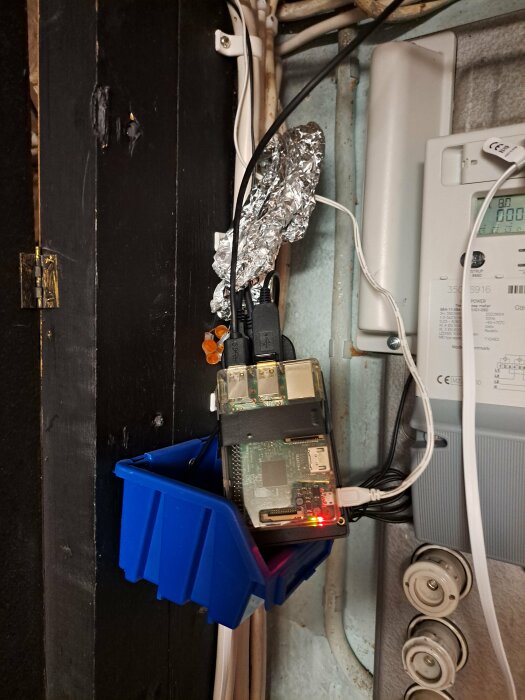 Improviserad elektronisk apparat, troligen Raspberry Pi, monterad på vägg, folie ovanpå, kablar, eluttag, rör nära.