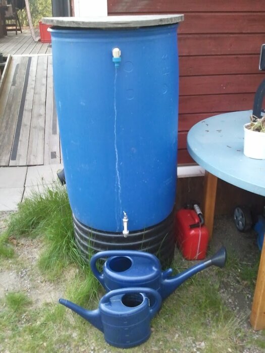 Blå regnvattentunna med kran, två vattenkannor, röd bensindunk, trädäck och hus i bakgrunden.