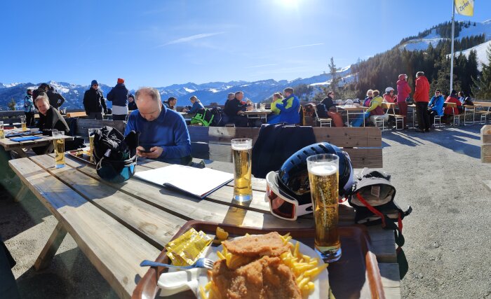 Skidåkare äter på bergsrestaurang, soligt väder, folk i vintersportkläder, snötäckta berg i bakgrunden.