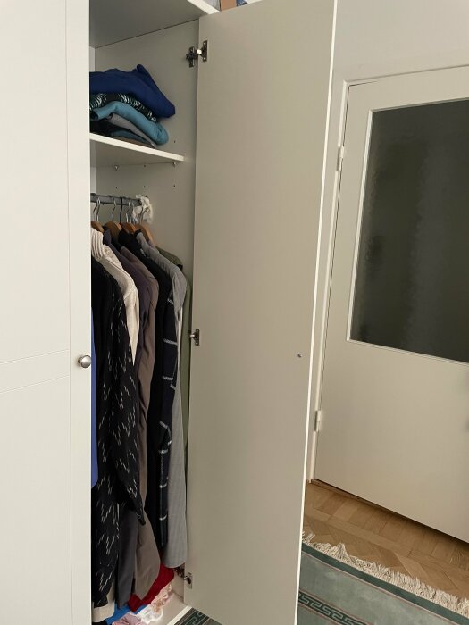 Öppen garderob med kläder på galgar och hyllor, matta och en dörr i ett rum.