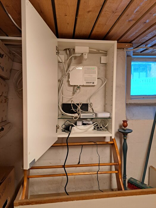 Öppet skåp med trådlös router, sladdar och teknisk utrustning ovanför en trähylla i ett rum.