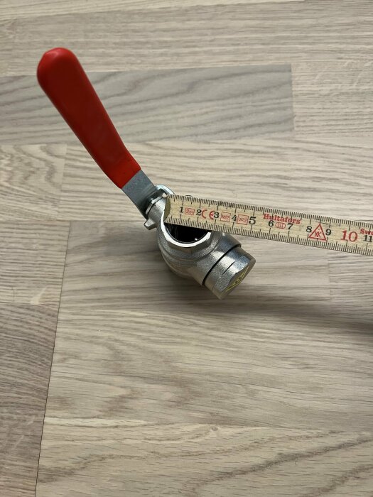 Ett måttband med rött handtag på ett träfärgat golv. Visar centimeter och tum. Redskap för mätning.