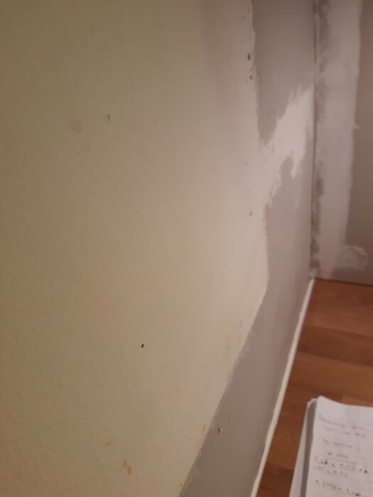 Hörn av rum med ojämn vitmålad vägg och golvlist; skrivblock synligt nertill. Renovering behövs möjligen.