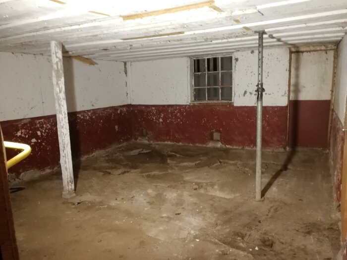 Förfallen källarlokal med smutsigt golv, stödpelare, vit och röd vägg, och ett litet fönster.