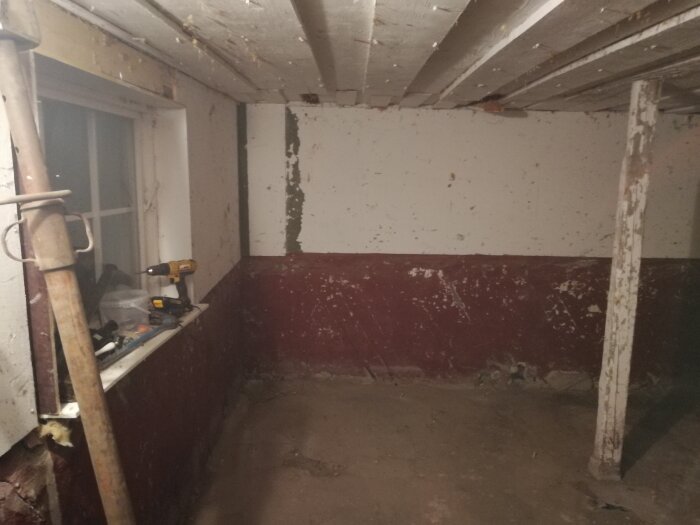 Ett slitet källarrum med vit och röd vägg, verktyg, fönster och synlig rördel.