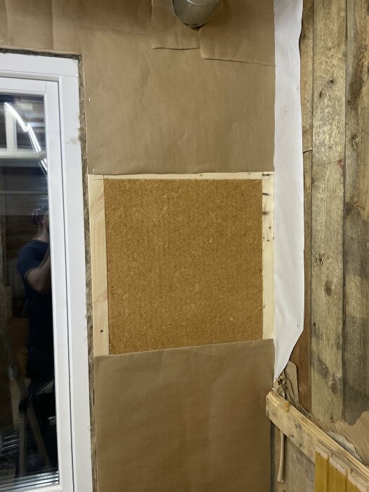 Inomhusrenovering, vägg med brunt papper och korkpanel, ofärdigt projekt, reflektion av person i fönster.