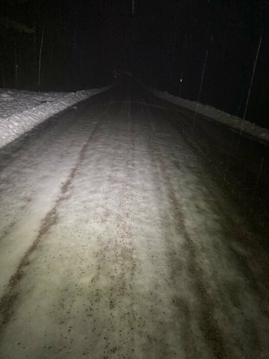 Mörk vinterväg omgiven av snö, upplyst av strålkastare, i nattmiljö.