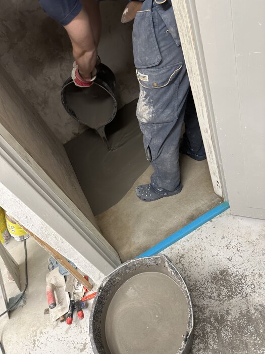 Häller nyligen blandad byggcement för att jämna ut golv inomhus; byggverktyg syns nära.