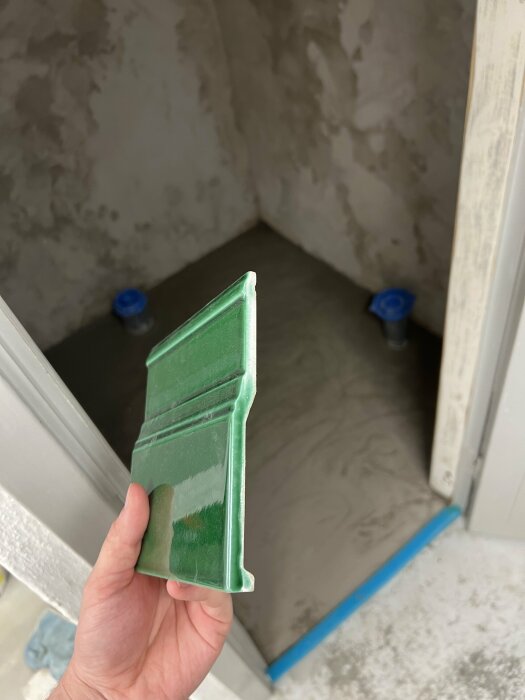 Hand håller grönt kakelprov framför halvfärdigt duschutrymme med synliga rör och spackel på väggarna.