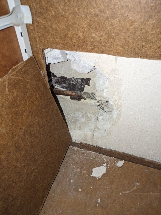 En skadad vägg med exponerade rör och isolering, nära en upphängningsskena och brunt golv, tecken på vattenskada eller renovering.