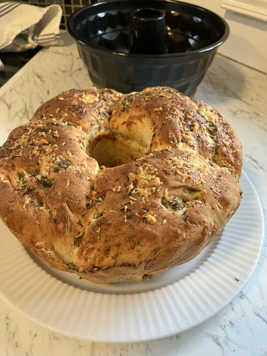 Nybakat kryddat bröd på vit tallrik, bundt-panna i bakgrunden, köksmiljö.