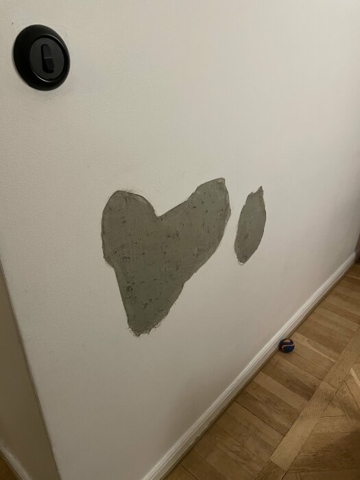 Vit vägg med hjärtformad spackling, svart dörrhandtag, trägolv, blå-turkos sandal nertill.