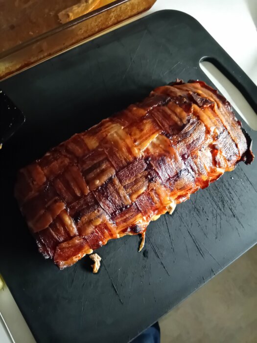 Ett baconlindat matbröd på en svart skärbräda, mycket gyllenbrunt och krispigt.