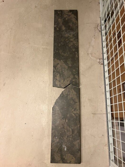Två stycken mörka kakelplattor på ett betonggolv, den ena är trasig.