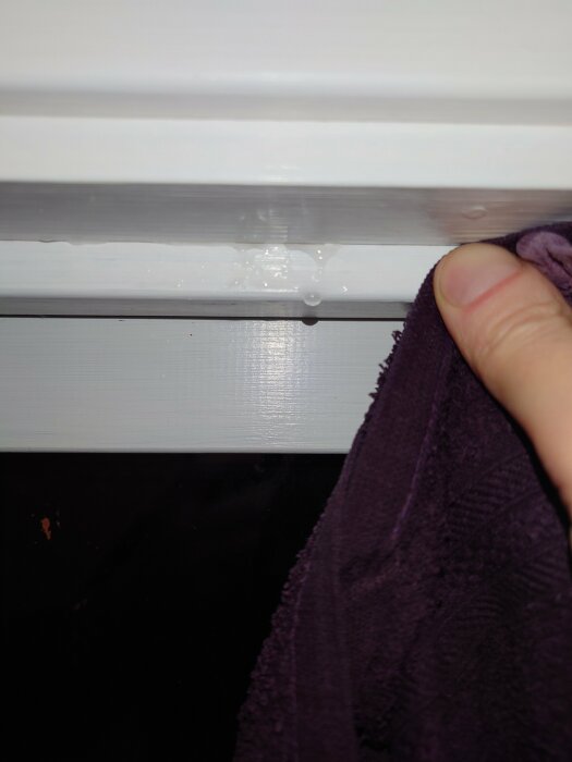 En hand med en lila trasa torkar kondens från en vit fönsterkarm.