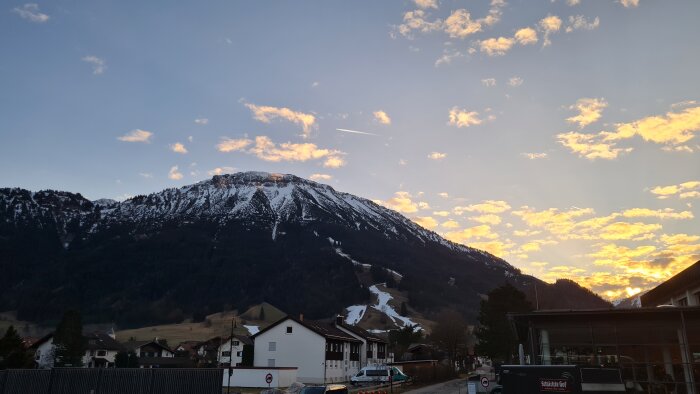 Solnedgång över snötäckt berg, himmel med orange moln, hus i förgrunden, lugnt och vackert landskap.