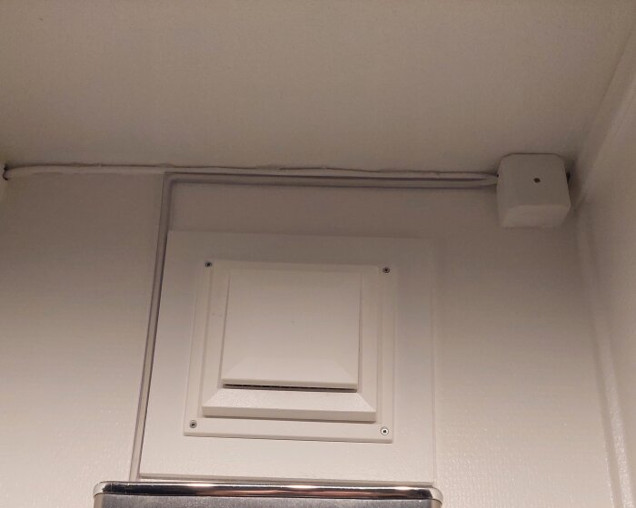 Ventilationslucka på vägg, säkerhetsdetektor i hörn, inomhus, sparsam belysning.