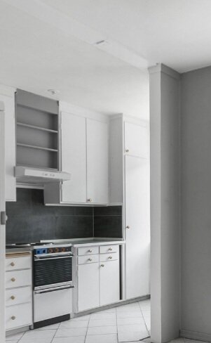 Ett hörn av ett minimalistiskt kök med vita skåp, svart arbetsbänk, spis och kaklat golv.