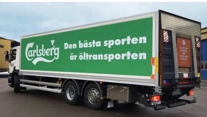 Lastbil med Carlsberg-logotyp, slogan om öltransport, parkerad utanför industribyggnad.