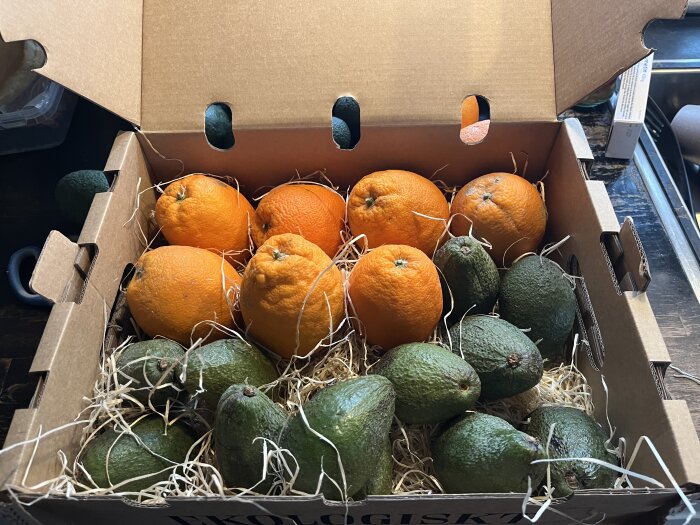 En kartong fylld med apelsiner och avokados, förpackat med stråliknande material, på ett köksbord.