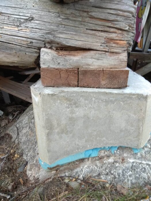 Bild av träbjälkar på betongblock, slitna, robusta, med blå isolering synlig vid grund. Construerad utomhusstödstruktur.
