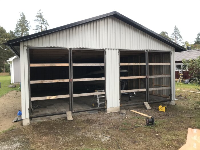 Delvis byggt garage med öppen framdel, träd och verktyg syns, grå himmel.