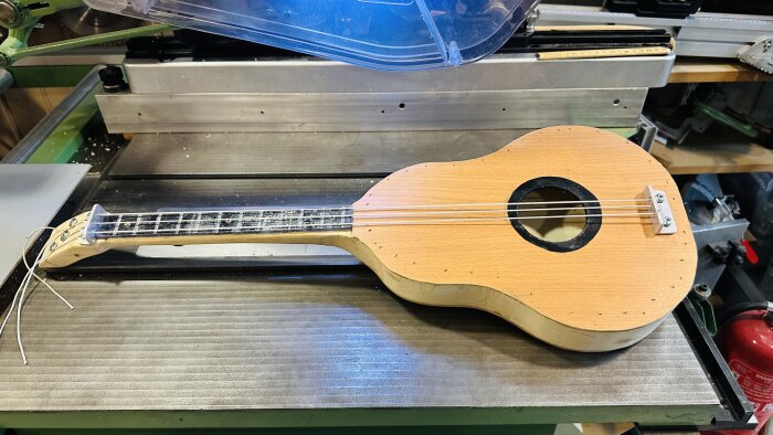 Obehandlad ukulele på en CNC-maskin i en verkstad, ofärdig, med spån och skärmekanism synlig.