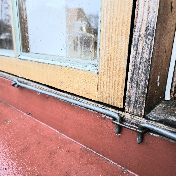 Närbild av fönsterkant med kondens, träets textur synlig, metallspärr i förgrunden, fuktig yta.