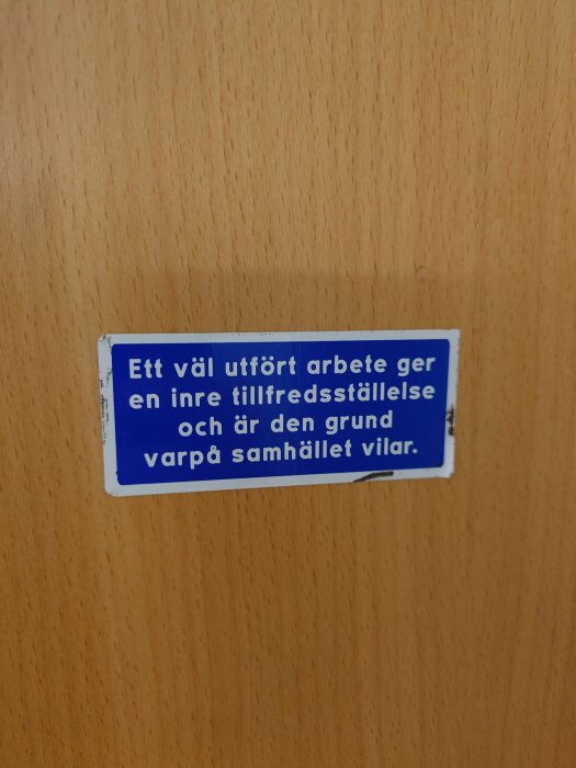 Blå etikett med text på träbakgrund: "Ett väl utfört arbete ger en inre tillfredsställelse och är den grund varpå samhället vilar."