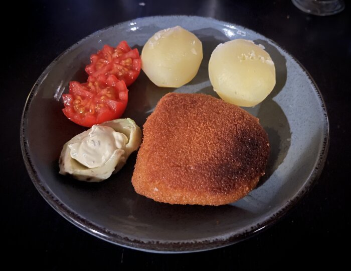 En tallrik med mat: panerad schnitzel, kokt potatis, artischockhjärta med majonnäs, skivad tomat. Enkel måltid.