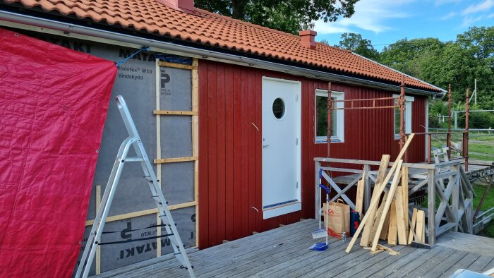 Röd stuga under renovering med byggmaterial och stegar på trädäck, omgivet av grönska.