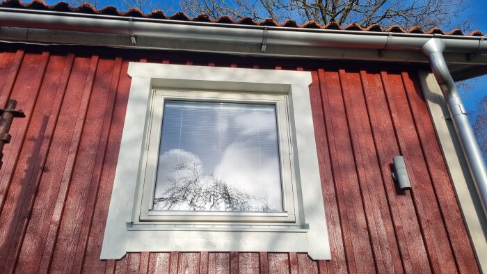 Ett fönster på en rödmålad trävägg med vita omfattningar under en blå himmel.