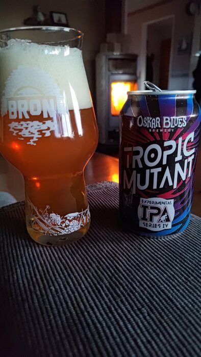 Ölglas med skum, burk från Oskar Blues Brewery, Tropic Mutant IPA, mysig bakgrund med brinnande kamin.