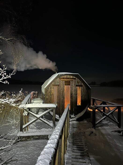 Vintervy med belyst bastu vid frusen sjö, ångmoln, snötäckt trädäck och brygga.