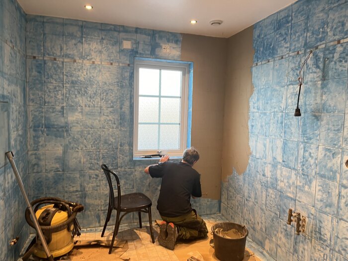 Renoveringsarbete pågår i ett badrum med en person som kaklar väggen. Verktyg och byggmaterial syns på golvet.
