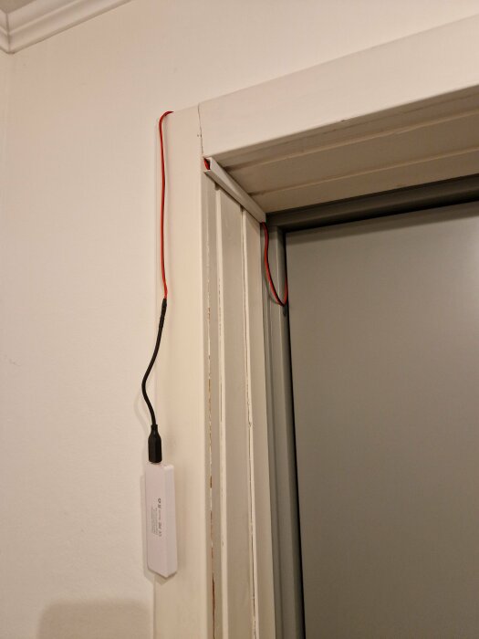 Röd och svart kabel löper längs en vägg och fönsterkarm med en elektronisk enhet.
