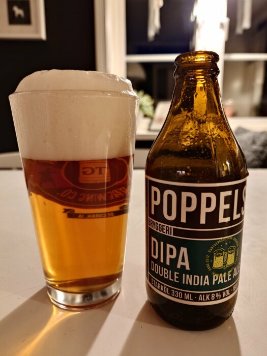 Ett glas med öl och en ölflaska märkt Poppels DIPA på ett bord framför fönster.