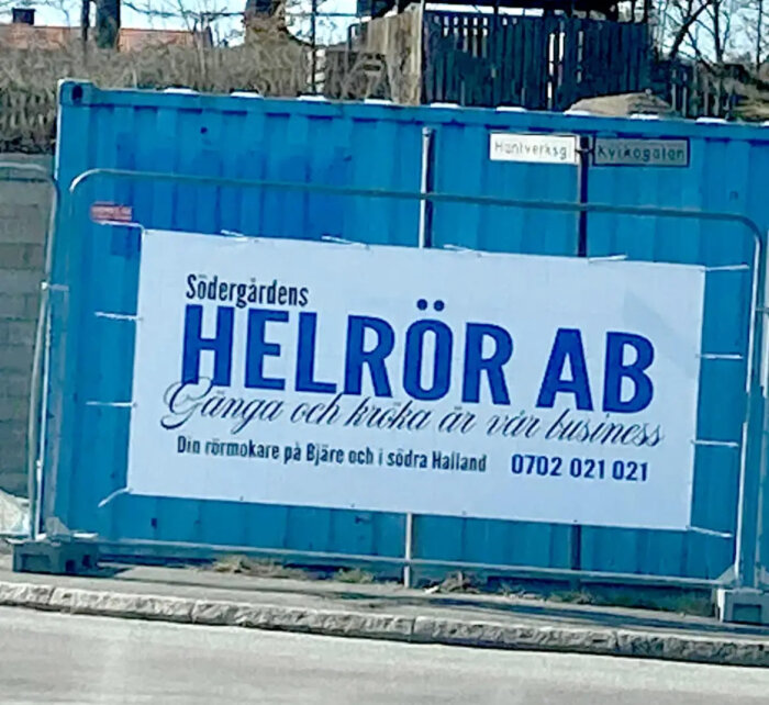 Blå container med vit banner, reklam för Södergårdens Helrör AB, kontaktinformation, slogan, gatunamnsskyltar ovanför.