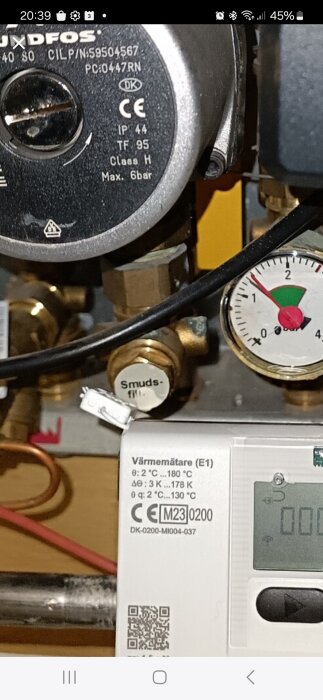 Cirkulationspump, manometer, rör och en värmeenergimätare i ett värmesystem. QR-kod, varningsikoner och tekniska data synliga.