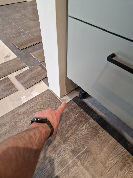 En hand pekar på en lucka vid golvsockel intill köksskåp, på två typer av golv.