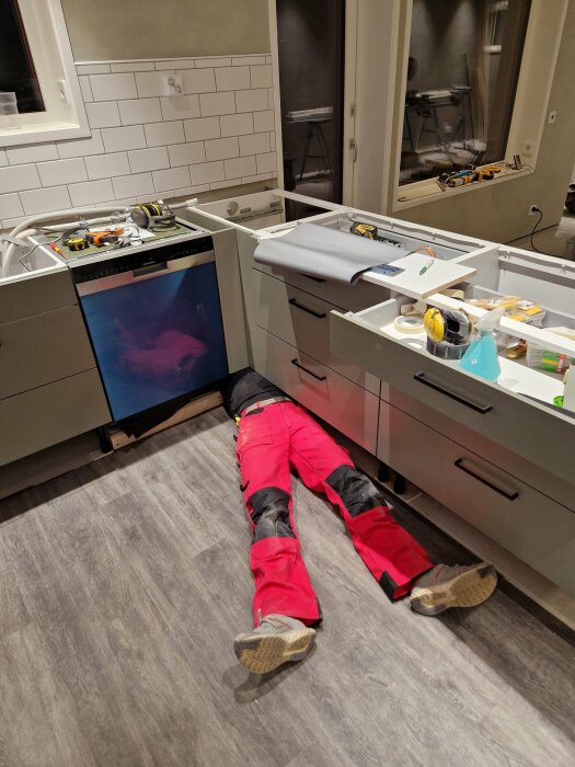 Omodernt kök under renovering med verktyg utspridda, person på golvet ger intryck av arbete bakom skåpluckor.