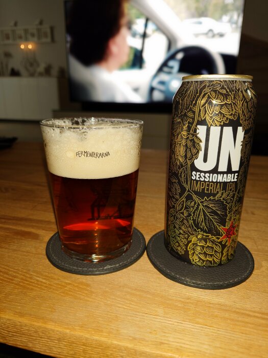 Ett glas öl och en burk IPA på bord framför suddig TV-skärm.