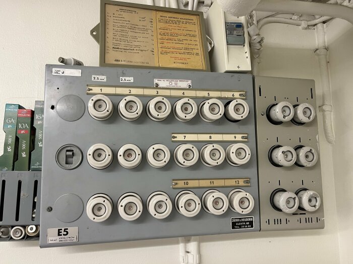 Äldre elektrisk säkringspanel med säkringar, märkningar, instruktioner, och knappar monterade på vägg.