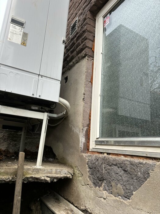 Utomhusvärmepump intill grovt putsad mur och fönster med mönstrad glasruta, över betonggrund och elledningar.