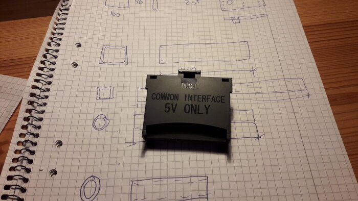 Elektronikmodul märkt "COMMON INTERFACE 5V ONLY" ovanpå rutigt papper med tekniska skisser, på träbord.