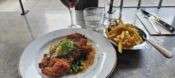 Panerad schnitzel med ärter, smör, sås, pommes frites, rödvin, vattenglas, bestick, marmorbord på restaurang.