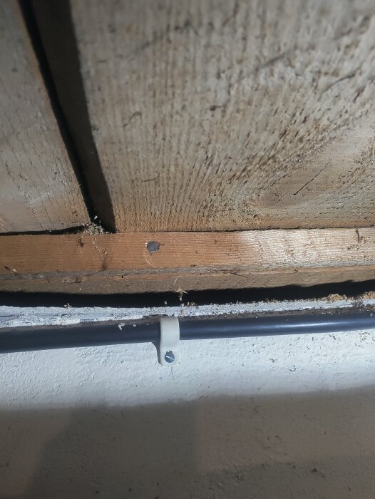 Bild på ett svart rör fäst vid en träbjälke med clips, dammigt, inomhus, oavslutade byggmaterial, skuggor.