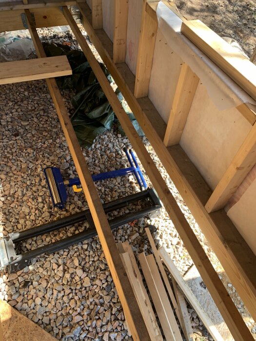 Byggprojekt utomhus visar trätrall, verktyg, sågbockar och material på grusunderlag.